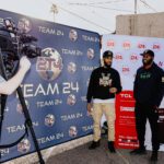 Two men standing in front of a wall with the team 2 4 logo on it.