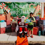 A boy standing next to santa clause in front of some presents.