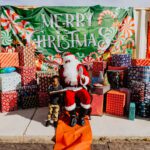 A santa clause sitting on the ground with many presents behind him.