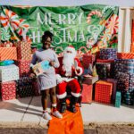 A woman standing next to santa clause in front of some presents.