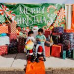 Two children sitting on a scooter in front of a merry christmas sign.