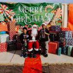 Two kids posing with santa claus in front of a merry christmas sign.