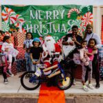 A group of people standing around a santa clause on a bike.